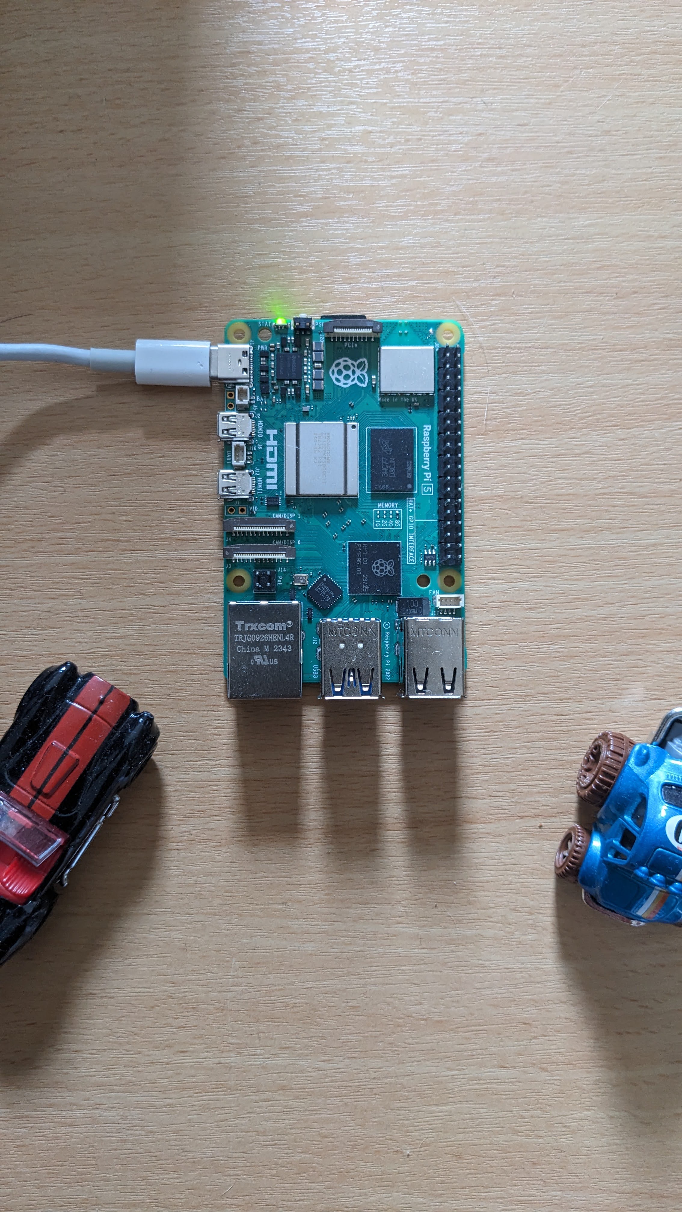 A Raspberry Pi 5 on a wooden desk with a USB-C power cable plugged in, flanked by two toy cars.